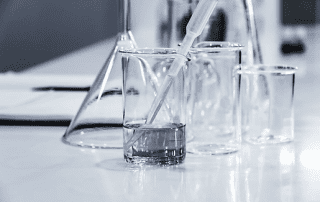 Laboratory glassware including beakers, flasks, and a pipette, partially filled with clear liquid, arranged on a reflective laboratory surface.