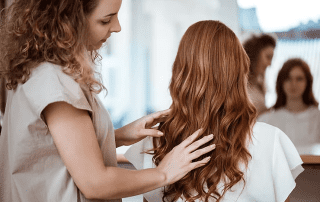 A hairstylist working on a woman's long curly hair in a salon, both reflected in a mirror in the background.