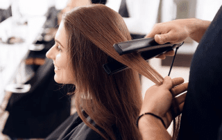 A woman smiling while a hairstylist uses a flat iron to straighten her long brown hair in a salon.