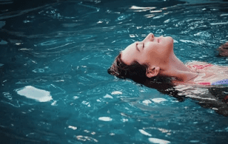 A woman floats peacefully on her back in a tranquil blue pool, her head slightly submerged and her face calm, surrounded by gently rippling water.