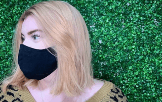 A blonde woman with a black face mask turns her head to the side against a lush green leafy background. she wears a leopard print sweater.