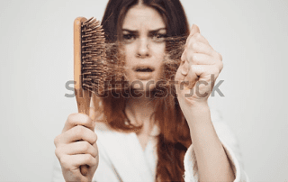 Young woman with a concerned expression holding a hairbrush with strands of hair in it, illustrating hair loss, against a plain background.