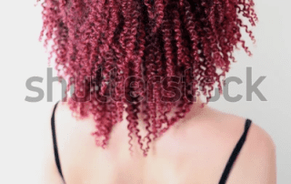 A woman with her back to the camera, showcasing vibrant red curly hair that covers her face against a light background. she wears a dark strap top.