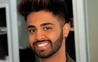 A young man with a stylish quiff hairstyle and a beard smiling at the camera. he wears a green jacket and an earring, set in an indoor environment with soft lighting.