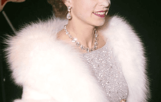 An elegant woman in a white fur coat and sparkling gown, adorned with a pearl necklace and earrings, smiles gently under a striped canopy. she holds a small golden clutch.