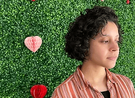 A profile of a person with short curly hair, wearing an orange striped shirt, standing in front of a green wall adorned with various red heart decorations.