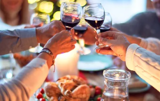 Group of friends toasting with red wine at a festive dinner table decorated with candles and a blurred christmas tree in the background.