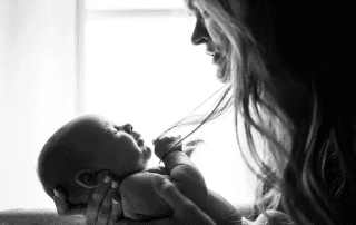 A black and white photo of a smiling woman holding and looking lovingly at a newborn baby in her arms, with a bright light background.