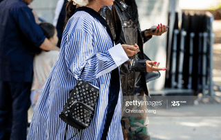 Two women converse outdoors, one dressed in a striped blue and white dress with a black purse, and the other in a camouflage outfit with sunglasses.