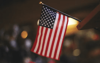 American flag hanging off the edge of a wooden surface, partially illuminated by a soft, warm light against a blurred background.