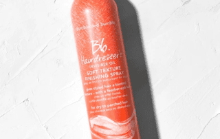 A bottle of bumble and bumble hairdresser's invisible oil soft texture finishing spray lying on a textured white surface, with sunlight casting a shadow on it.