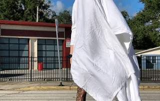 A person wearing a ghost costume made from a white sheet with eye cut-outs stands on a sunny street, revealing fishnet stockings and black boots.