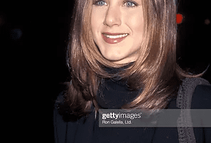 A woman with shoulder-length straight brown hair and a black coat smiles gently at a public event. the background features blurred lights.