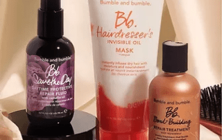 A collection of bumble and bumble hair care products displayed against a draped beige fabric background. items include sprays, a mask, and a treatment oil, all in stylish packaging.