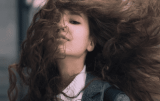 A young woman with long, voluminous curly hair blowing wildly around her face as she tilts her head back, accentuating a playful expression. she's wearing a cozy sweater and a jacket.