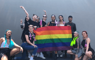 A diverse group of people joyfully posing with a large rainbow pride flag at an outdoor event. they are smiling and dressed in colorful, expressive outfits, showing a vibrant sense of community and celebration.