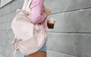 A woman in a pink jacket and denim shorts, holding an iced coffee, with a pink backpack over her shoulder, stands against a gray wall.