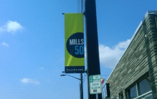 A bright yellow banner with "mills 50" in large black letters, hung on a street pole under a clear blue sky, showcasing the mills 50 district. link (mills50.org) at the bottom.