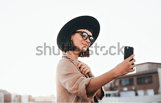 A stylish young woman with a black hat and glasses takes a selfie with her smartphone on a sunny rooftop. she is smiling and dressed in a casual beige jacket.