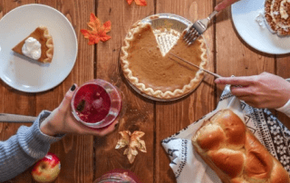 A cozy autumnal meal setting featuring hands cutting into a creamy pumpkin pie, a cup of cranberry sauce held aloft, challah bread, an apple, and plates with desserts on a wooden table decorated with fall leaves.