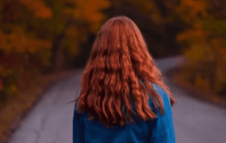 A person with long, wavy red hair viewed from behind, standing on a winding road surrounded by vibrant autumn foliage.
.