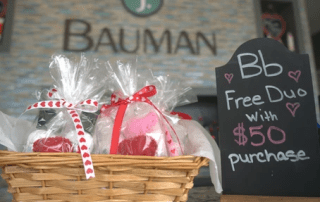 A wicker basket filled with wrapped gifts sits on a counter with a chalkboard sign offering a free duo with $50 purchase, background features a sign reading "bauman.