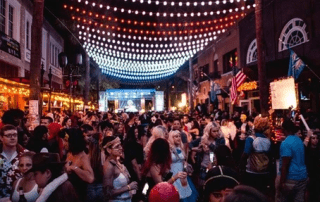 A busy outdoor street party at night, decorated with strings of overhead lights, and crowded with people in various costumes, suggesting a festive or carnival atmosphere.