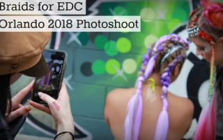 A person captures a photo of two women with colorful braided hairstyles at a photoshoot for edc orlando 2018, with a blurred background featuring lights.