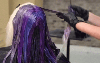 A hairstylist wearing black gloves dyes a customer's hair with vibrant purple and blue colors, focusing on applying color to the back section of the hair.