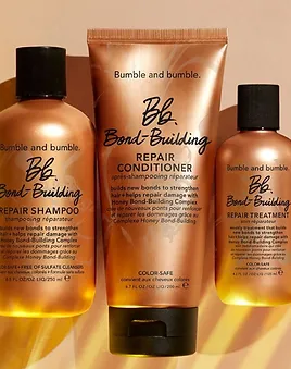 Three bumble and bumble hair care products on a warm background: bond-building repair shampoo, conditioner, and repair treatment, labeled in bronze and white.