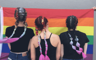 Three women with long, braided hair standing in front of a rainbow flag, holding its edges. each braid is adorned with brightly colored ribbons.