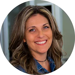 Portrait of a smiling middle-aged woman with wavy brown hair, wearing a denim jacket, in a circular frame with a blurred background.