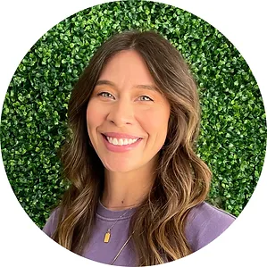 A smiling woman with long brown hair wearing a purple shirt stands in front of a lush green foliage background, forming a circular frame around her.