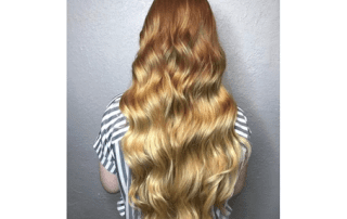 A woman's back view showing her long, wavy hair colored in a gradient that transitions from auburn at the roots to blonde at the tips. she wears a black and white striped shirt.