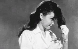 A black-and-white gif of a young woman with long hair, wearing a white blouse, playfully twirling her hair with her finger and smiling coyly.