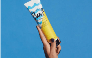 A person holds a tube of "surf" branded sunscreen against a solid blue background. the hand is visible with purple nail polish on the fingers.