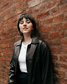 A young woman with dark hair and bangs stands confidently in front of a brick wall, wearing a black leather jacket over a white top.