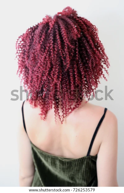 A woman with her back to the camera, featuring vibrant red curly hair, wearing a dark green spaghetti strap top against a white background.