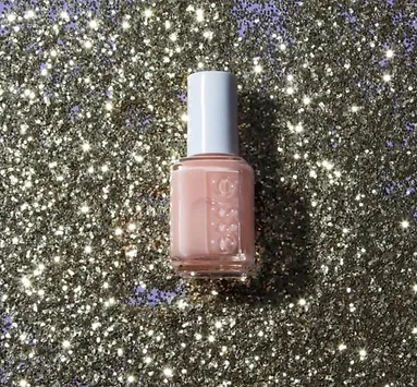 A bottle of light pink nail polish stands centered on a glittering gold background that sparkles brightly in the light.