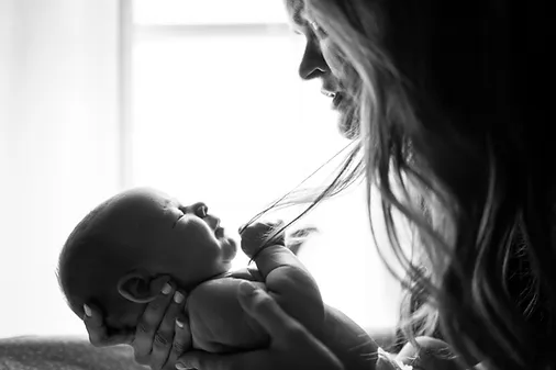 Black and white image of a joyful mother holding her newborn baby, looking at each other affectionately, with soft light streaming in from the background.