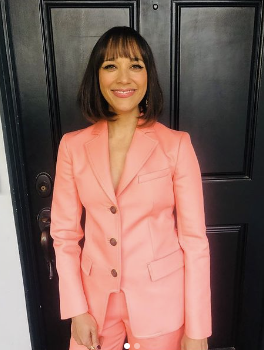 A woman with shoulder-length brown hair, smiling, dressed in a coral pink blazer and trousers, standing in front of a black door.