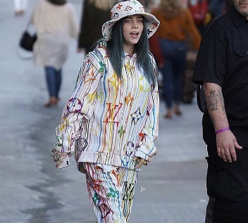 Billie eilish wearing a colorful, patterned outfit with a bucket hat, walking on a street with people walking behind her. her hair is dyed green and blue.