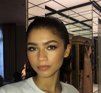 A close-up portrait of a young woman with sleek, pulled-back hair and striking makeup, featuring prominent eyelashes and glossy lips, in a room with mirrored walls.