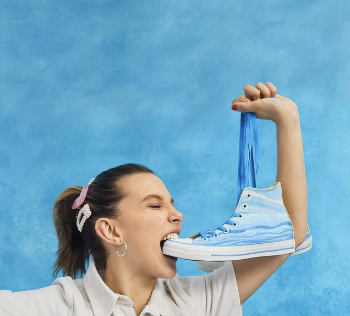 A playful image of a woman biting a blue sneaker with paint dripping from it, against a light blue background. she is wearing a white shirt and has her hair in a ponytail.