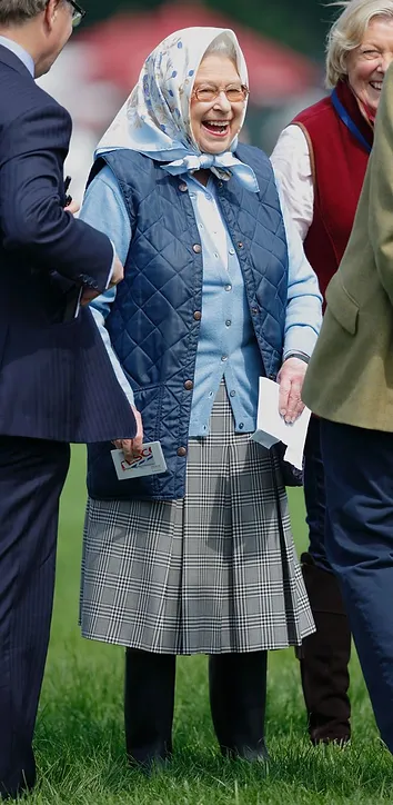 An elderly woman in a blue vest, white blouse, and plaid skirt wearing glasses and a patterned headscarf smiles broadly while walking outdoors with two other people.