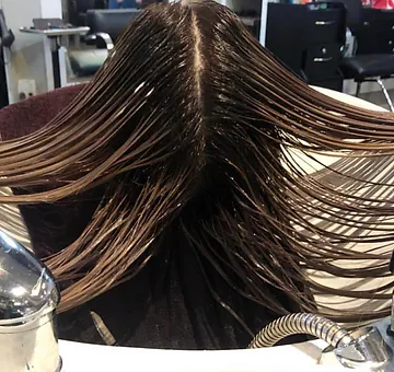 A woman's head is draped forward over a salon sink, showing her splayed wet hair, highlighted brown, during a hair wash or treatment. salon environment visible in the background.