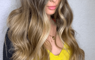 A woman with long, wavy hair featuring blonde highlights, wearing a yellow top and a delicate necklace, posing with a partial view of her face visible.