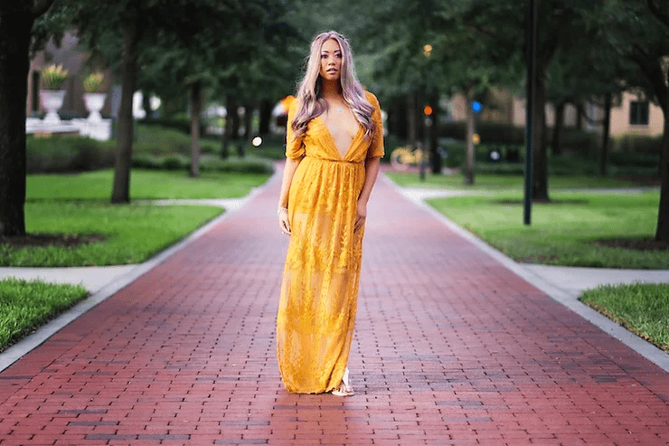 A woman in a flowing yellow dress stands confidently on a brick pathway, surrounded by green grass and trees, with lamp posts softly glowing in the background at dusk.