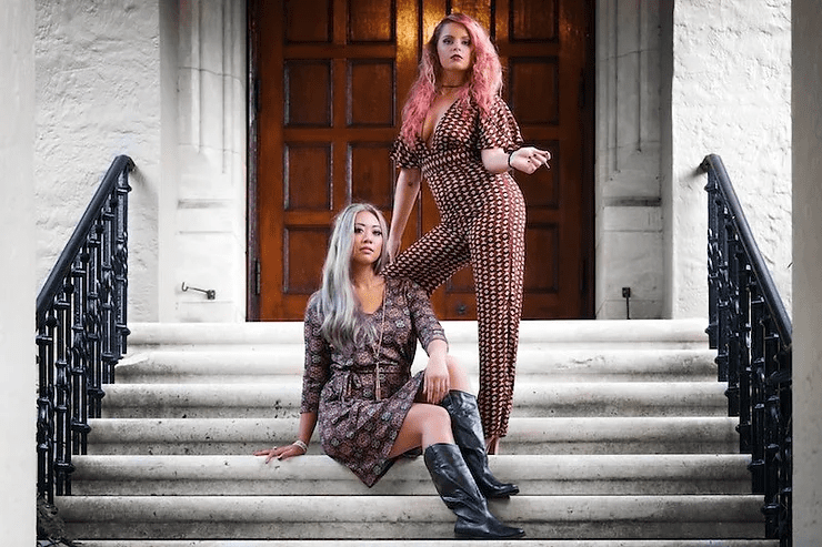 Two women posing on stone steps; one sits with long silver hair, a patterned dress and black boots, while the other stands with vibrant red hair and a matching patterned jumpsuit.