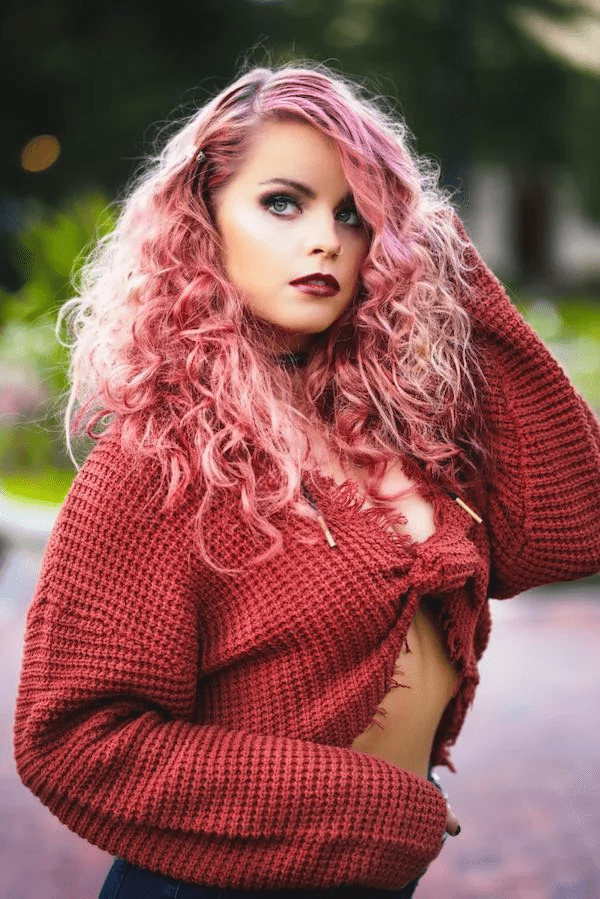 A woman with curly pink hair and bold makeup poses outdoors, wearing a chunky, rust-colored sweater draped off one shoulder. lush greenery softly blurs in the background.